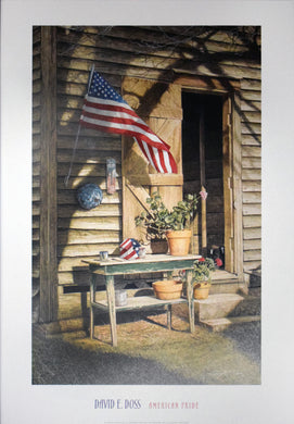 David E Doss American Pride  18 by 26 inchesposter with white border. Image of a cabin with the flag on the outside blowing in the wind above a table of plants resting against the house. There is a birdhouse with an american flag painted on it resting on the green table and the door to the homestead is open revealing a key. 