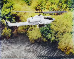 AH-1W Super Cobra Helicopter flies above water in photograph with green trees next to the water the helicopter is white and says marines on it it leaves ripples in the water as it flies away 